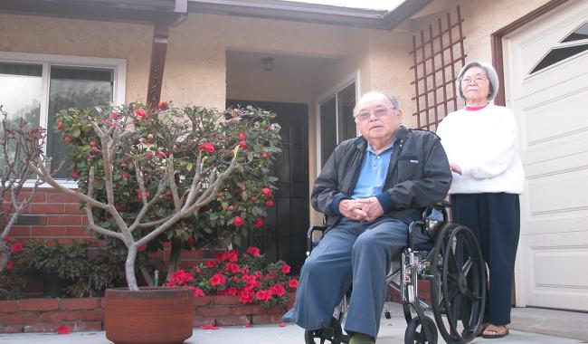 A senior woman standing by an elderly man in a wheelchair in front of a single-family home