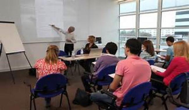 An image of classroom showing adults sitting at desk facing a teacher point at a board